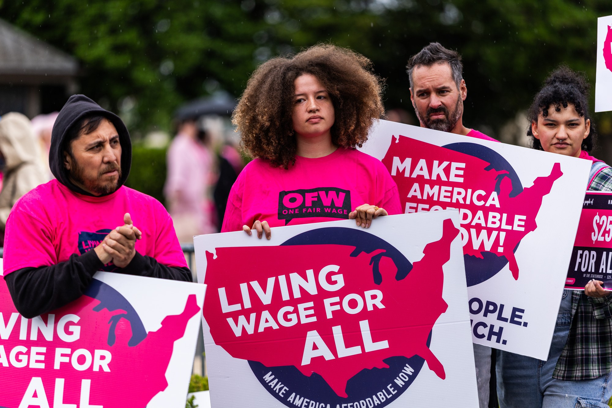 Four OFW worker members in pink shirts holding Living Wage For All and Make America Affordable Now signs at a rally