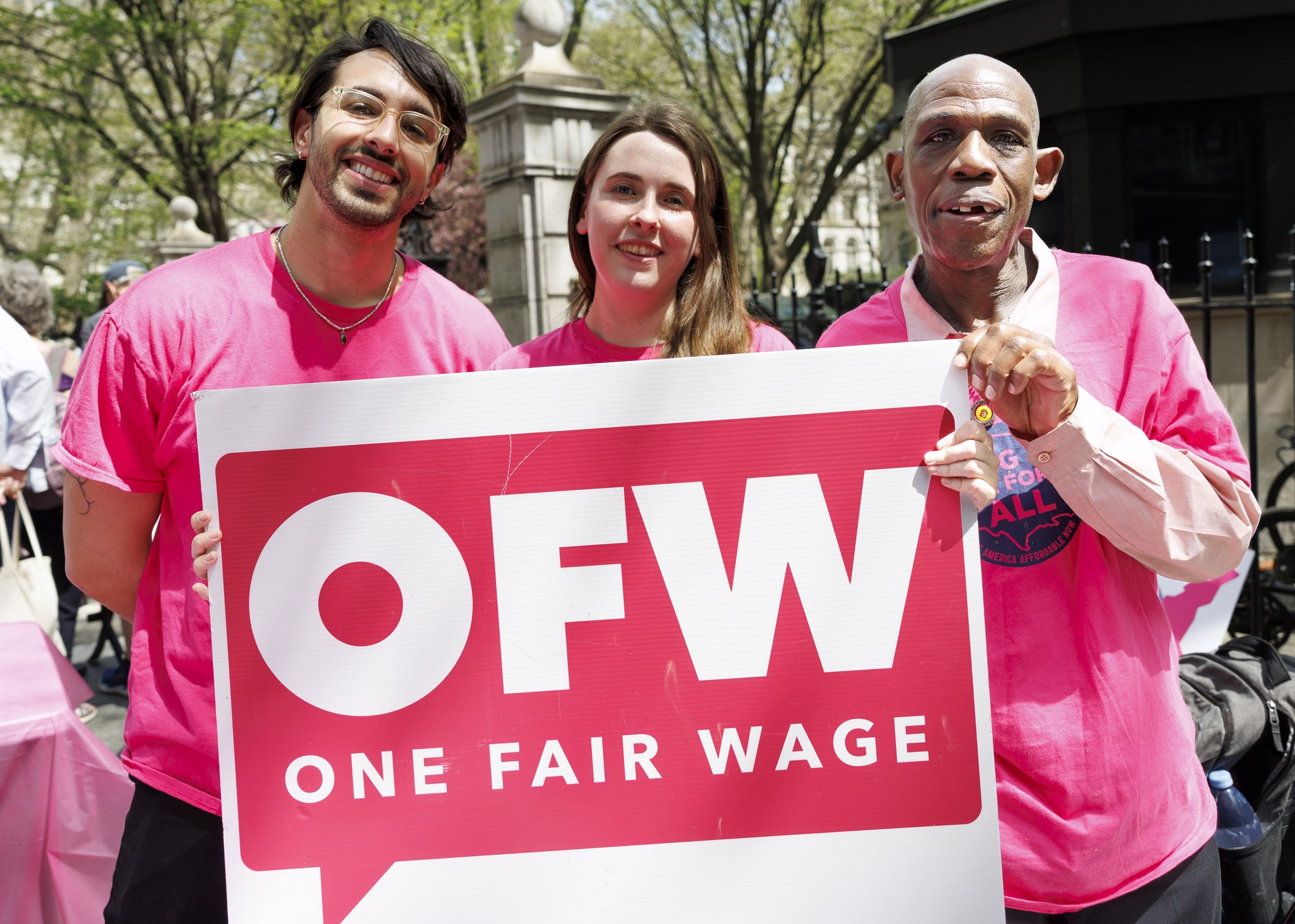 OFW members and staff in pink shirts hold the OFW logo speech-bubble sign at the NYC City Hall action, April 16, 2026